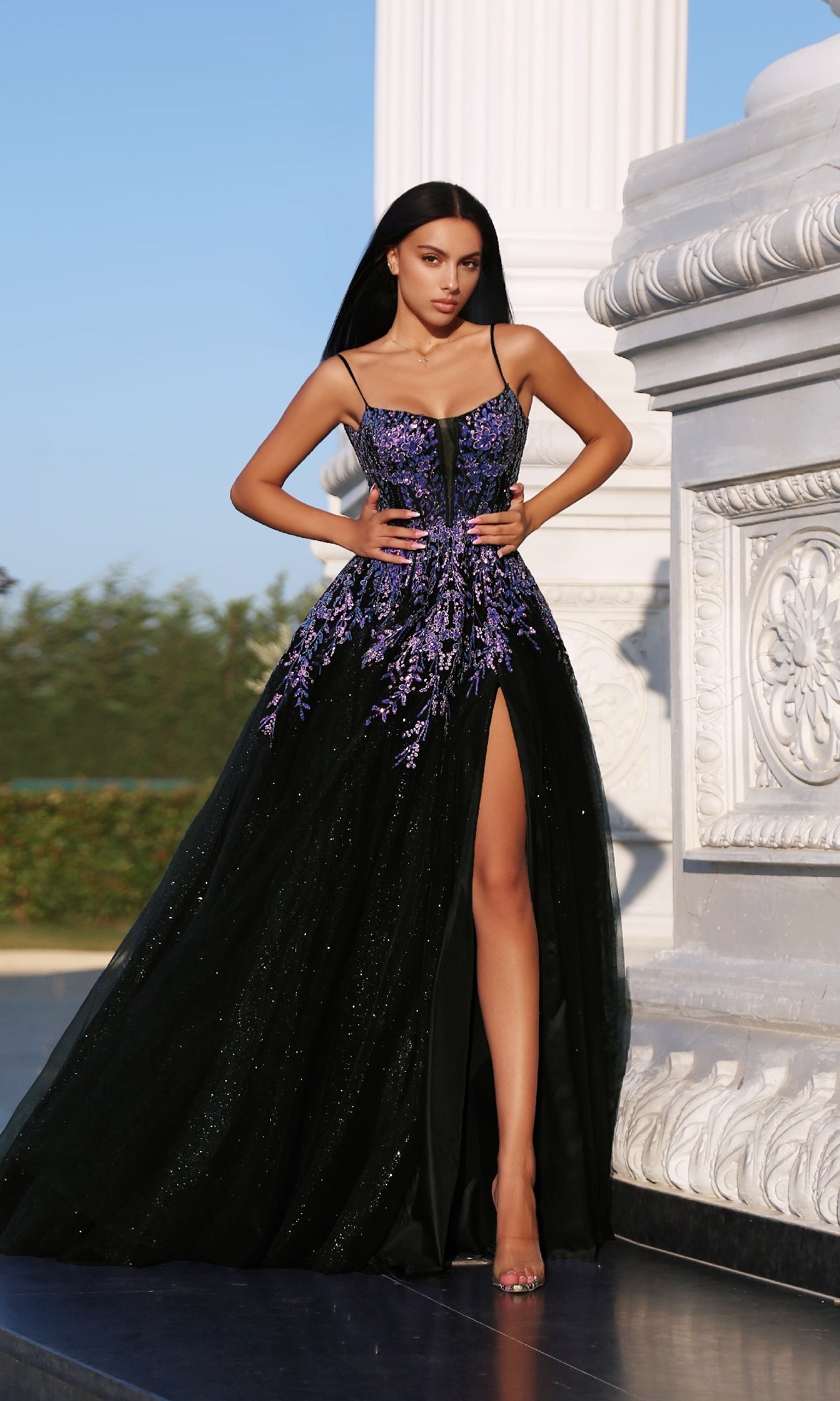 A woman stands outdoors beside a white ornate structure, wearing the Tina Holly TE216B Long Formal Dress—an elegant black and purple evening gown with floral appliques and a high-leg slit.